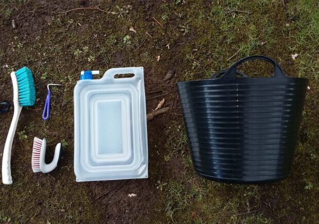 Equipment for a biosecurity kit is in a line on the ground outside. From left to right there is a brush, bootpick, water container and bucket