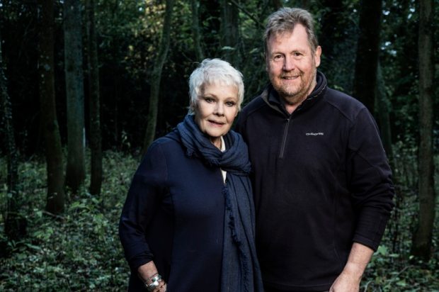 Judi Dench stands to the left of the image looking at the camera and Tony Kirkham stands next to her on the right also looking at the camera. They are central in the image