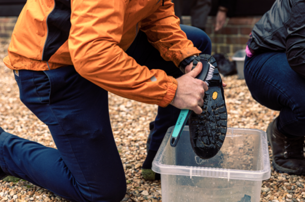 A person wearking jeans and an orange raincoat kneels down on one knee beside a large plastic box holding a boot upside down over a box, which they are scrubbing with a brush held in the other hand.