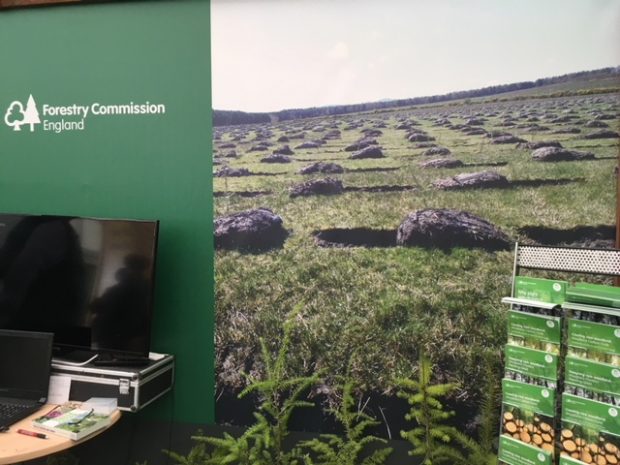 A computer sits to the left of the image in front of a large banner depicting lots of dug holes with the soil piled to the side of them and the words Forestry Commission England. A display of green leaflets sits to the right of the image