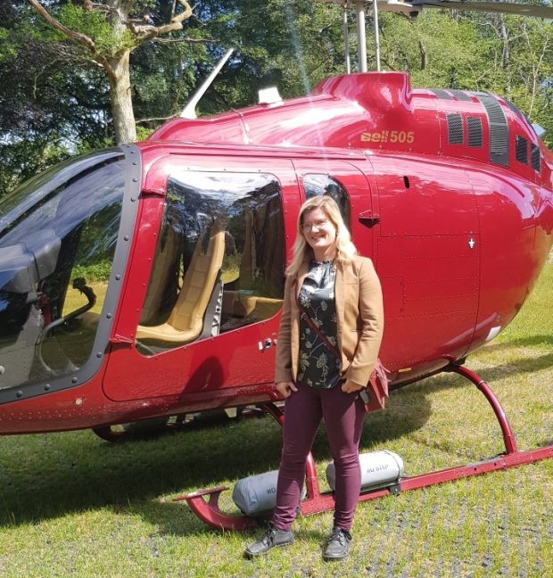 A woman with light brown hair and glasses stands in front of a red helicopter with her hands in her pocket