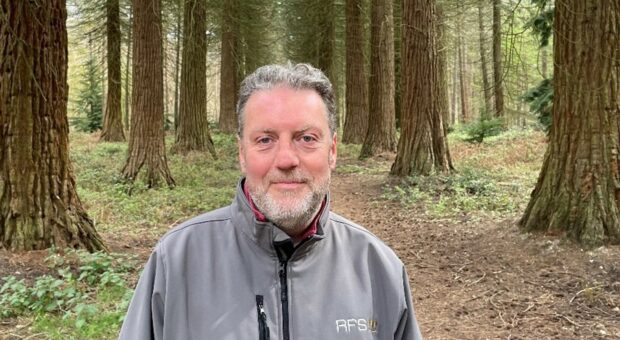 A man waring a grey jacket standing in woodland smiles at the camara