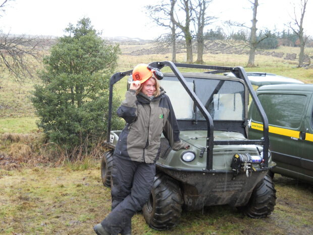 Kate leans against a vehicle parked in a field slightly lifting the hard hat on her head