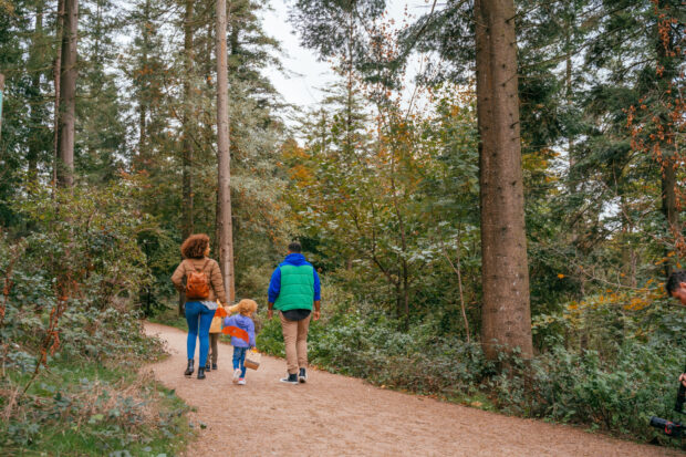 Two adults and two children walk along a forest path wearing warm clothing. One adult holds hands with one of the children