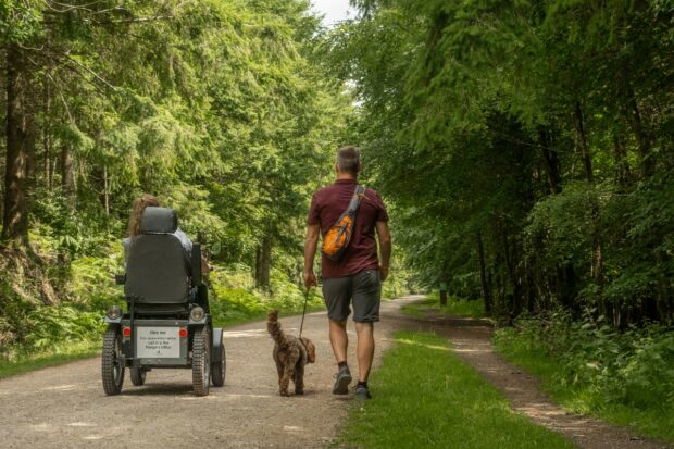 A person rides in a tramper along a woodland path next to a person walking a dog