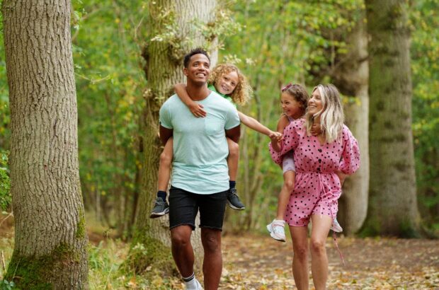 A family: a man, woman and two children on the adults back, walk through a woodland wearing summer clothes