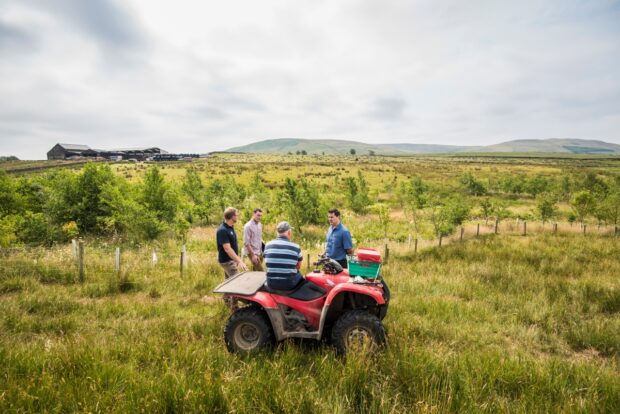 A person sitting on a quad bike facing three people standing up and talking to them. The quad bike is in a grassy field with trees in the background